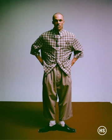 Man in oversized shirt and pants poses in studio with hands on hips, standing on brown floor against pale blue background, fashion shoot.