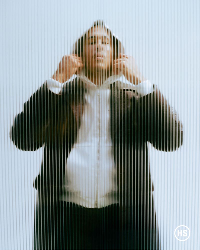 Fashion shot of a person wearing a white hoodie and black jacket, standing against a white background with black vertical stripes.