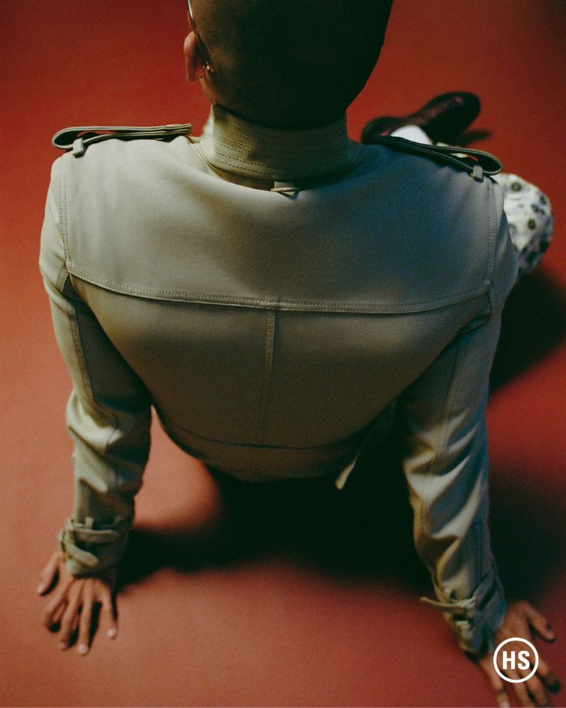 Person seated on red surface wearing a light tan military-style jacket with epaulettes in a top-down shot, showing back details.