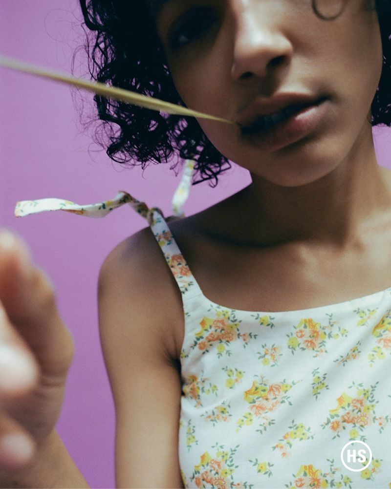 Close-up of a young woman with curly dark hair, wearing a floral top, holding a blade of grass in her mouth, against a purple background.