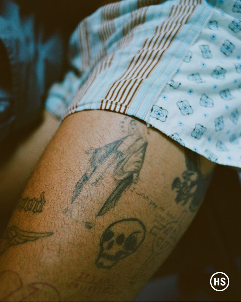 Tattooed arm close-up shows skull, sword and other tattoo art on fair skin, set against a blue patterned shirt sleeve.