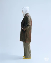 Model in a brown coat and white hoodie standing against a light grey studio background