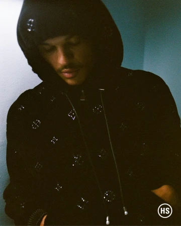 Model wearing a black hooded jacket with monogram pattern under dim indoor lighting for Highsnobiety.