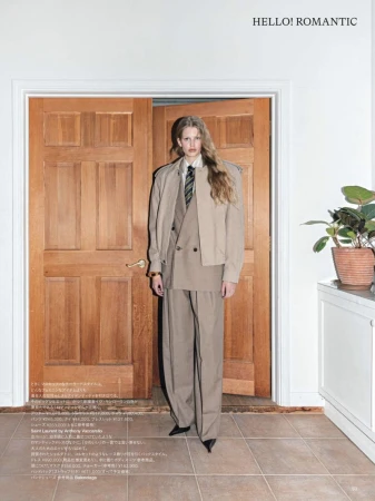 A fashion-forward woman in a beige suit stands in a doorway, casting a gaze in an indoor setting with a potted plant.