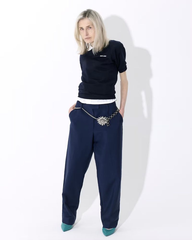 Fashion portrait of a blonde adult woman in a dark sweater, blue pants with a silver chain belt on a white background.