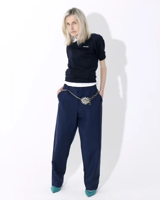 Fashion portrait of a blonde adult woman in a dark sweater, blue pants with a silver chain belt on a white background.
