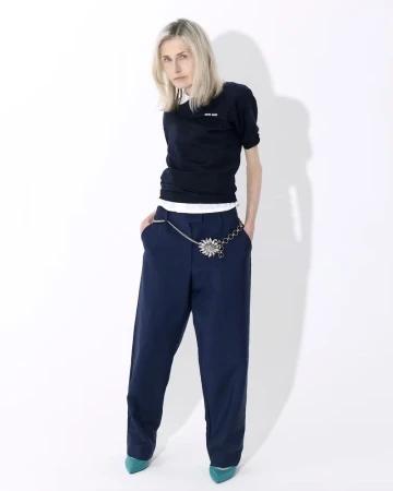 Fashion portrait of a blonde adult woman in a dark sweater, blue pants with a silver chain belt on a white background.