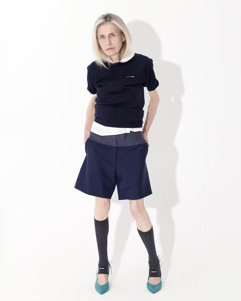 Full-length portrait of an adult woman with blonde hair, wearing a navy top, blue shorts, black socks, and teal high heels, against a white backdrop.