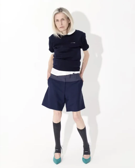 Full-length portrait of an adult woman with blonde hair, wearing a navy top, blue shorts, black socks, and teal high heels, against a white backdrop.