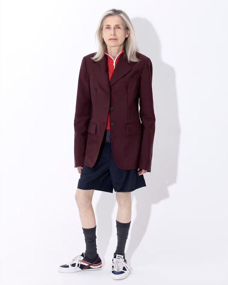 An older woman with long gray hair in a maroon blazer and navy shorts stands in a photo studio against a seamless white background.