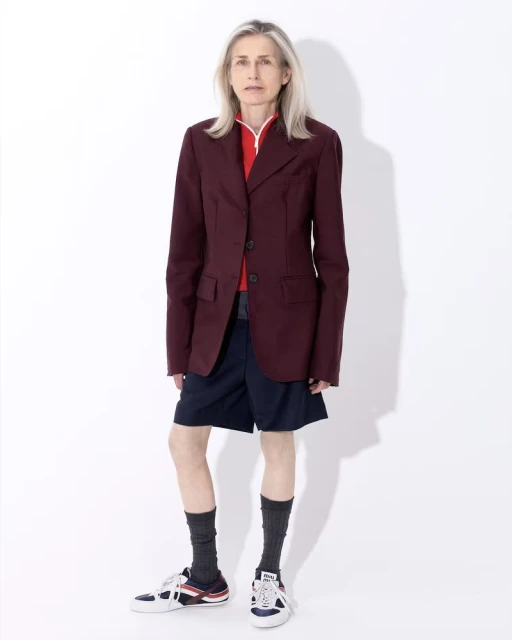 An older woman with long gray hair in a maroon blazer and navy shorts stands in a photo studio against a seamless white background.