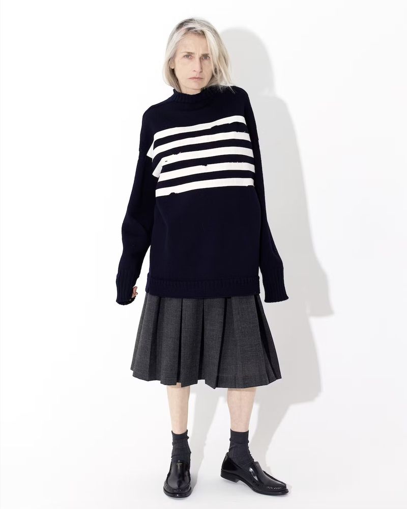 A woman with short gray hair stands in a photo studio wearing a blue and white striped sweater, pleated gray skirt, and shiny black shoes.