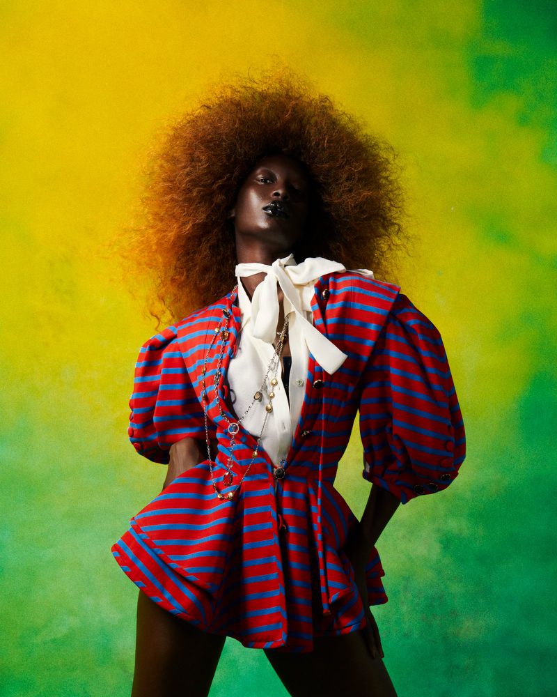 Fashion shot of a Black woman with afro in a red and blue striped playsuit in front of a yellow and green blended background.
