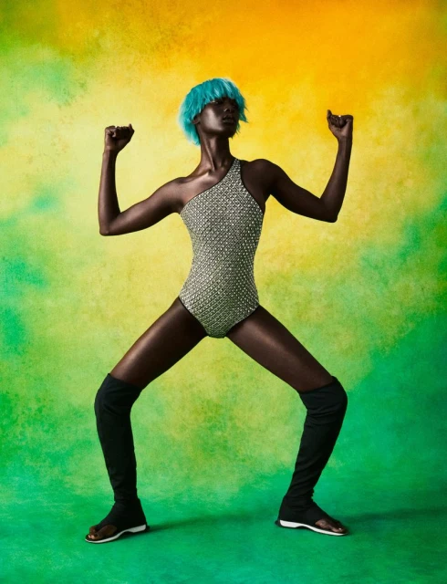 A slender Black woman with blue hair poses in a one-shoulder bathing suit with thigh-high socks against a painted yellow and green background.