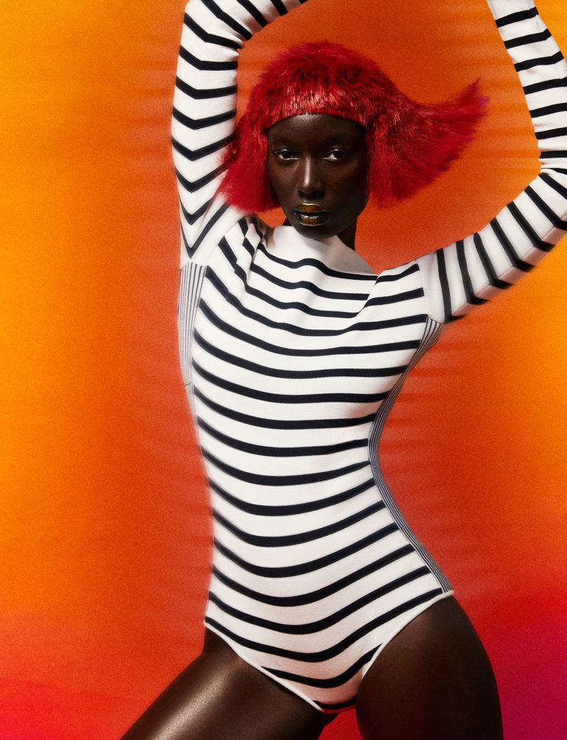 An African American woman with a short red wig, gold lipstick, and a striped bodysuit poses in front of an orange background.