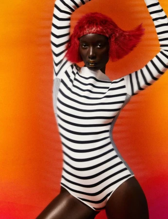 An African American woman with a short red wig, gold lipstick, and a striped bodysuit poses in front of an orange background.