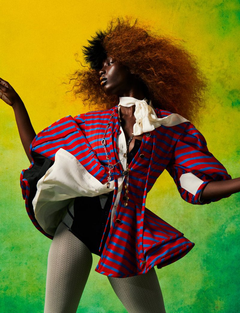 Fashionable woman with afro wearing a red and blue striped jacket with a white ruffled collar posing in front of a yellow and green background.