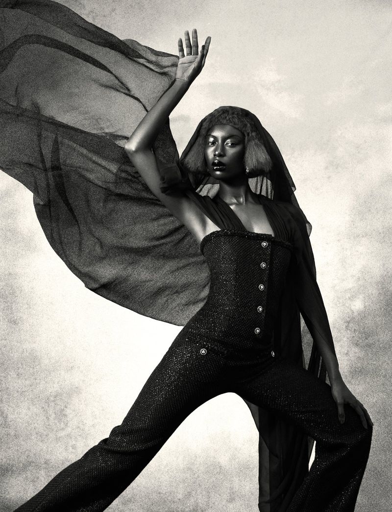A single African-American woman in a sparkling jumpsuit and flowing fabric poses dramatically against a mottled, neutral background in a studio shot.