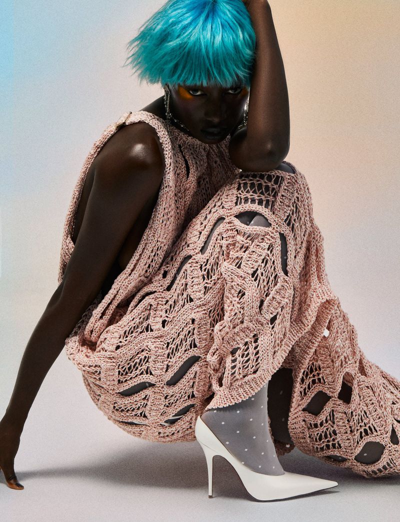 A Black female fashion model with blue hair poses indoors wearing a pink crocheted dress and white heels for a beauty and fashion shoot.