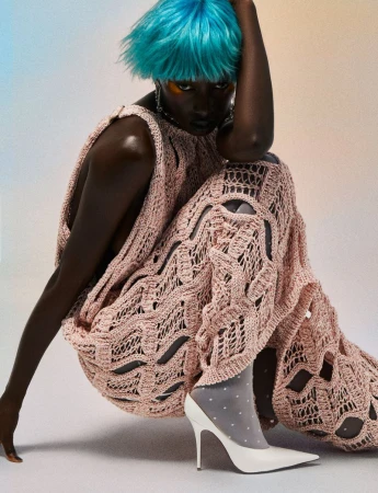 A Black female fashion model with blue hair poses indoors wearing a pink crocheted dress and white heels for a beauty and fashion shoot.