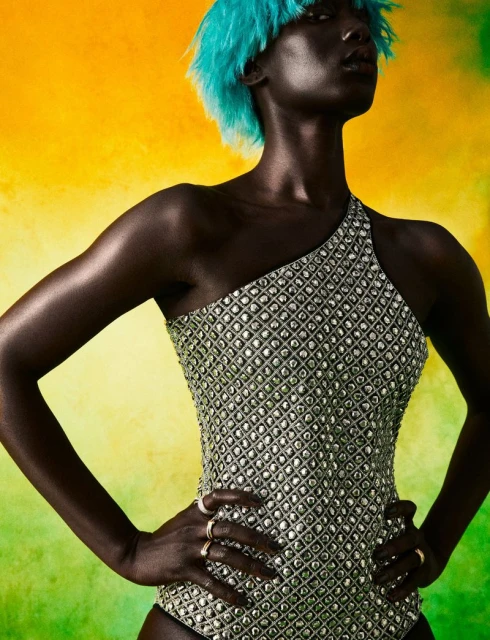 Close-up of a Black woman with aqua hair posing in a shimmering silver one-shoulder bodysuit against a vibrant yellow and green backdrop.