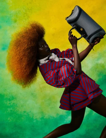 Model with voluminous red hair holding a blue handbag against a vibrant yellow and green watercolour wash background.