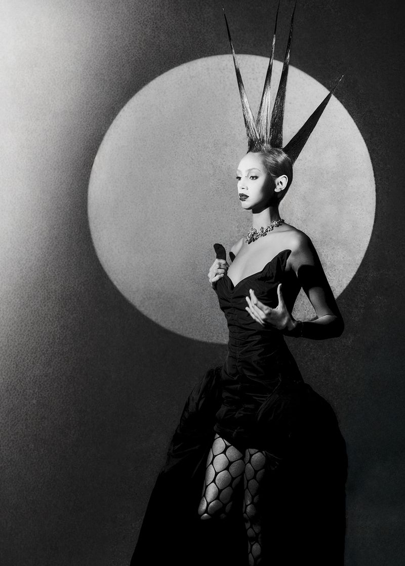 Model with avant-garde spiky hair wearing a black dress and patterned tights against a circular backlit backdrop.