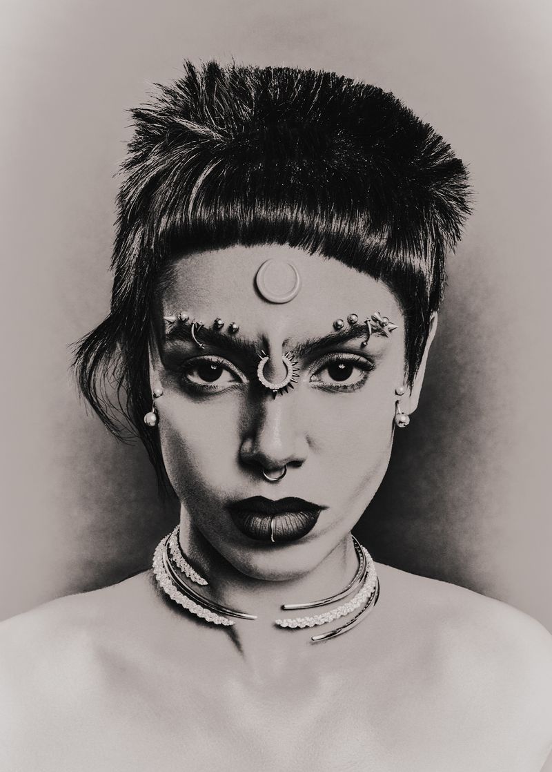 Monochrome portrait of a woman with striking facial piercings and dark lipstick, complemented by a unique hairstyle and pearl necklace.