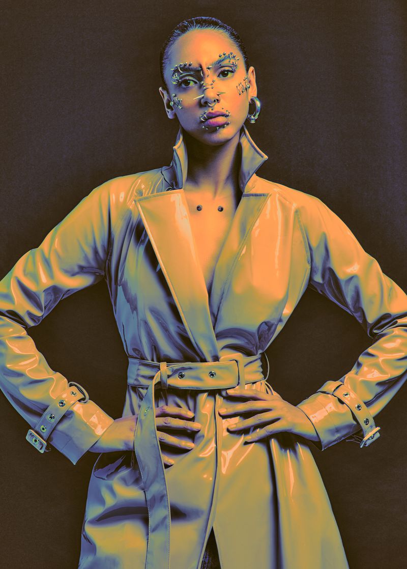 Woman in orange trench coat with hands on hips, posing against a dark background, wearing face jewelry and statement earrings.