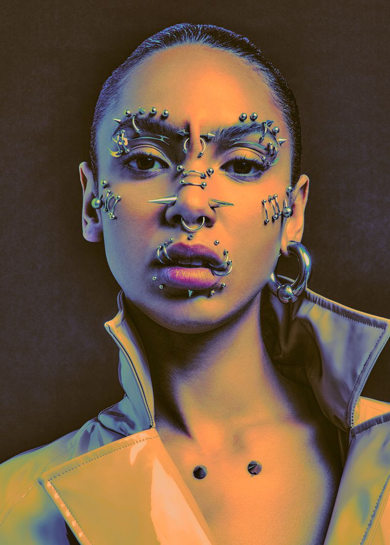 Headshot of an adult woman with numerous piercings wearing a shiny trench coat in front of a dark background, fashion portrait.