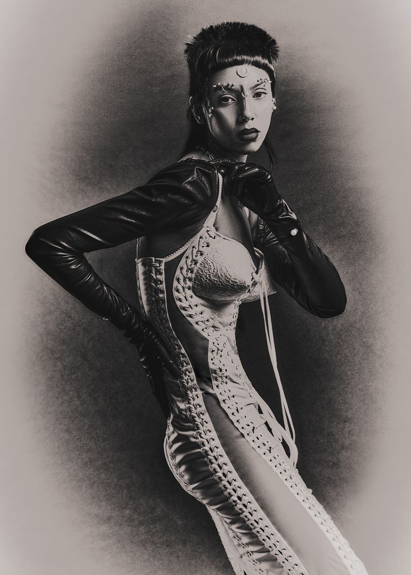 Model wearing a lace-up corset and long black leather gloves with artistic face makeup against a textured monochrome background.