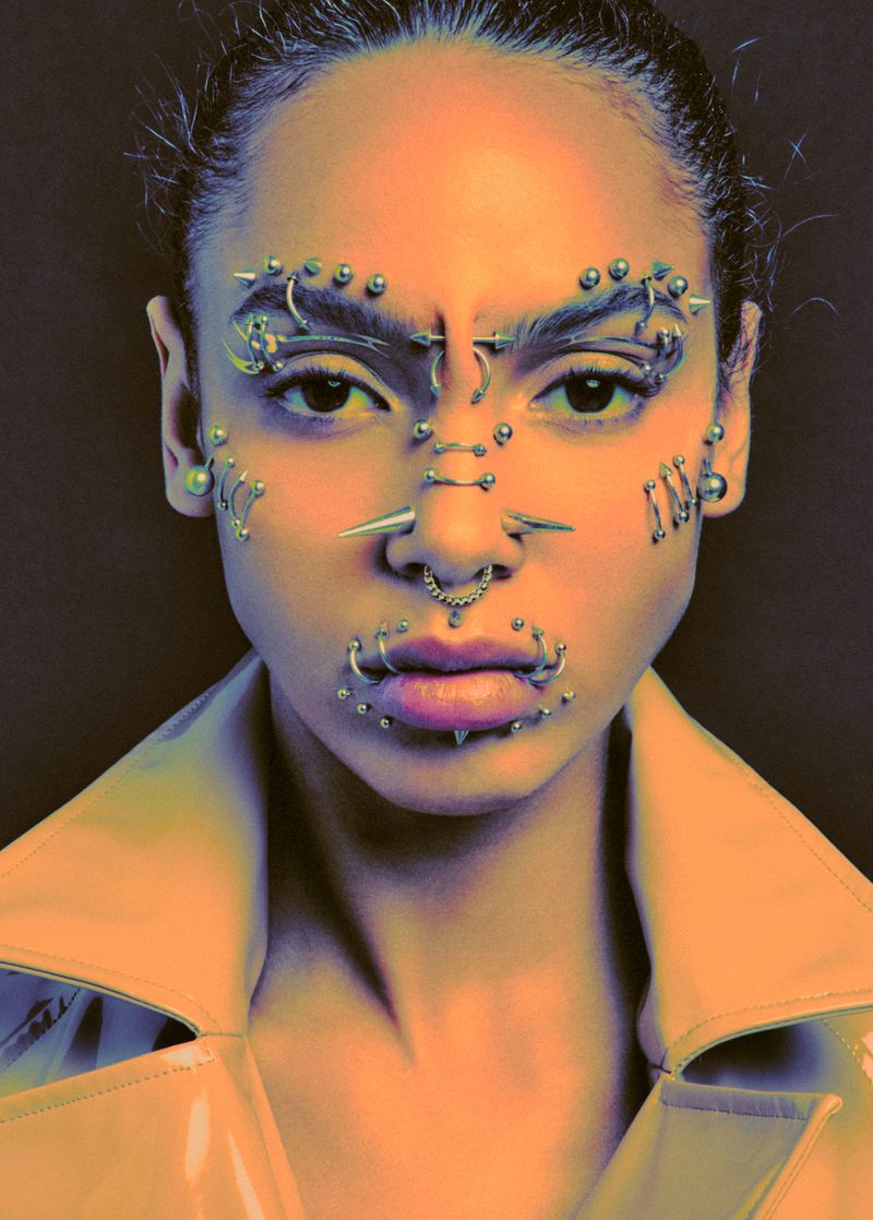 Portrait of an adult female with heavy facial piercings looking directly at the camera in a studio with a neutral dark background.