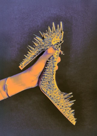 A hand holds a silver high-heeled shoe covered in spikes against a dark background in a fashion or beauty-related image.