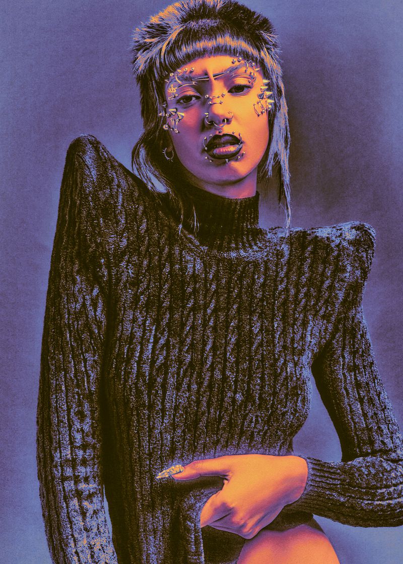 Portrait of adult female wearing a knit sweater, hat with fur trim, and facial jewelry, posing against a grey and violet backdrop.