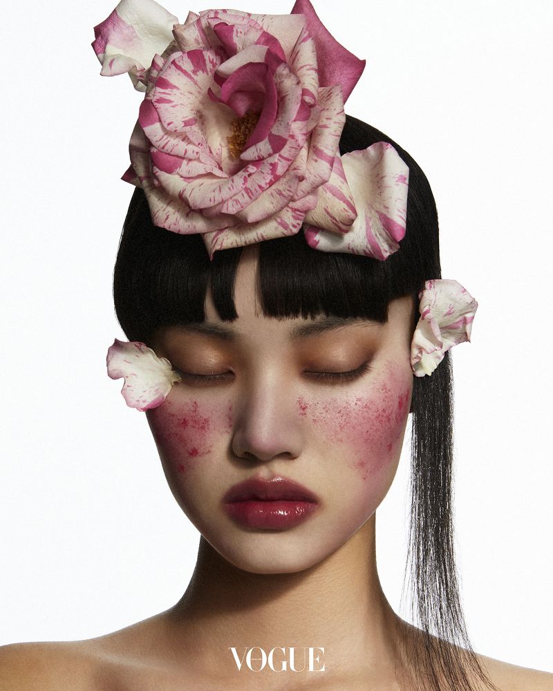 Close-up beauty shot of an Asian woman with closed eyes, wearing floral headpiece and pink blush, set against a bright white background.