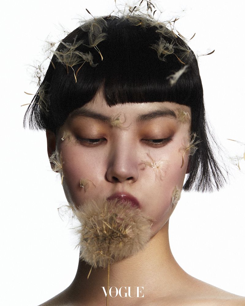 Close-up portrait of an Asian woman with short dark hair and light makeup, adorned with dandelion seeds, against a clean white backdrop.
