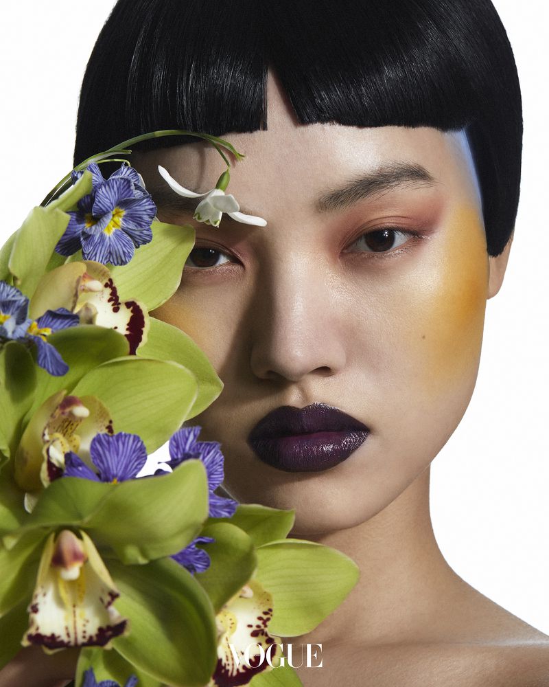 Portrait of an Asian woman with short black hair and dramatic makeup, set against a bright background with orchids and violets.
