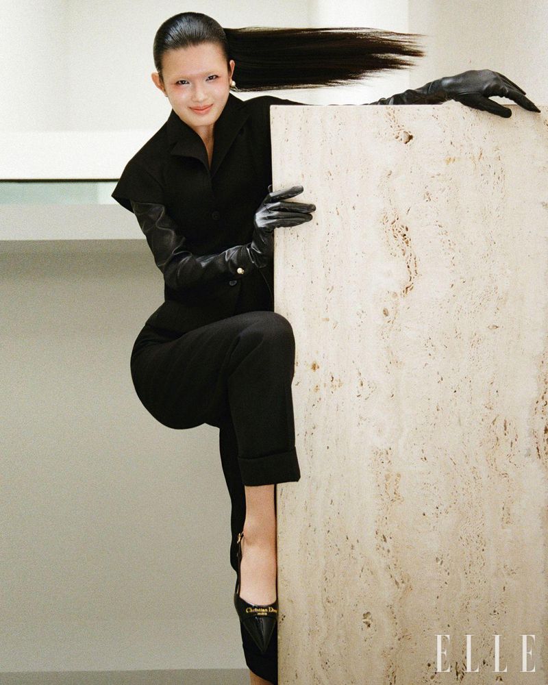 A woman in a black suit and gloves leans against a travertine slab, high ponytail, captured in a fashion editorial style shot with 'Elle' logo.