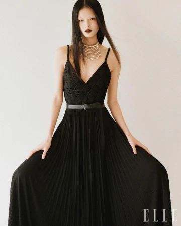 Model in a long black pleated evening dress with a thin belt and layered pearl necklace against a plain background