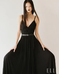 Model in a long black pleated evening dress with a thin belt and layered pearl necklace against a plain background