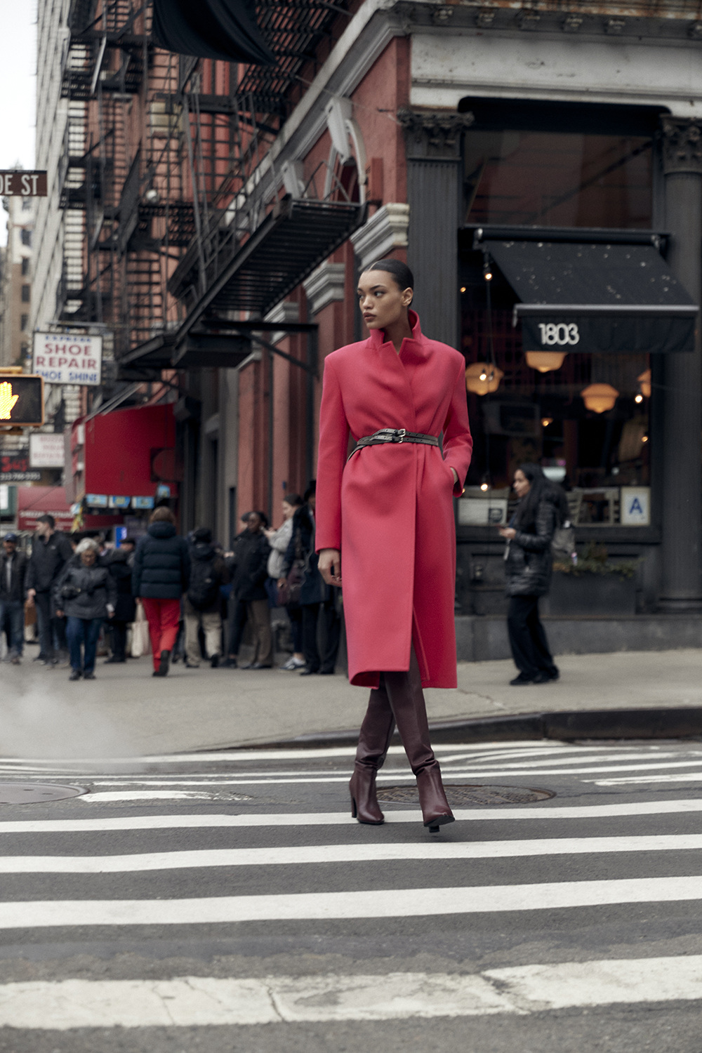 A fashionably dressed woman in a red coat crosses a zebra street, with city buildings and pedestrians visible in the background.