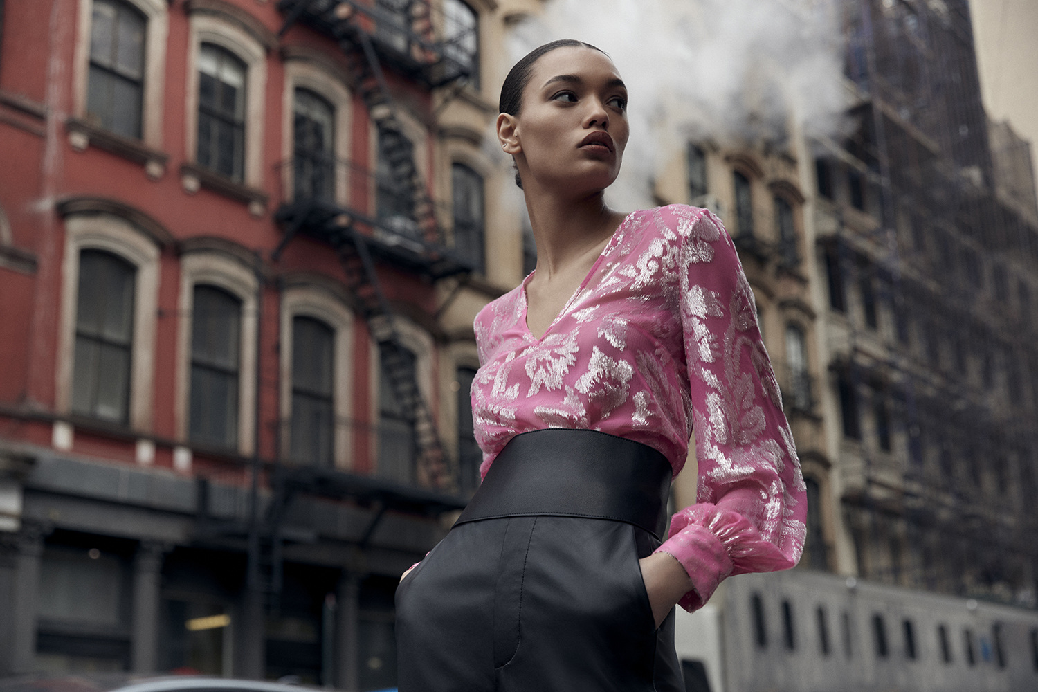 Adult woman in a pink floral top and black pants poses on a city street with red brick buildings and vapor in the background.
