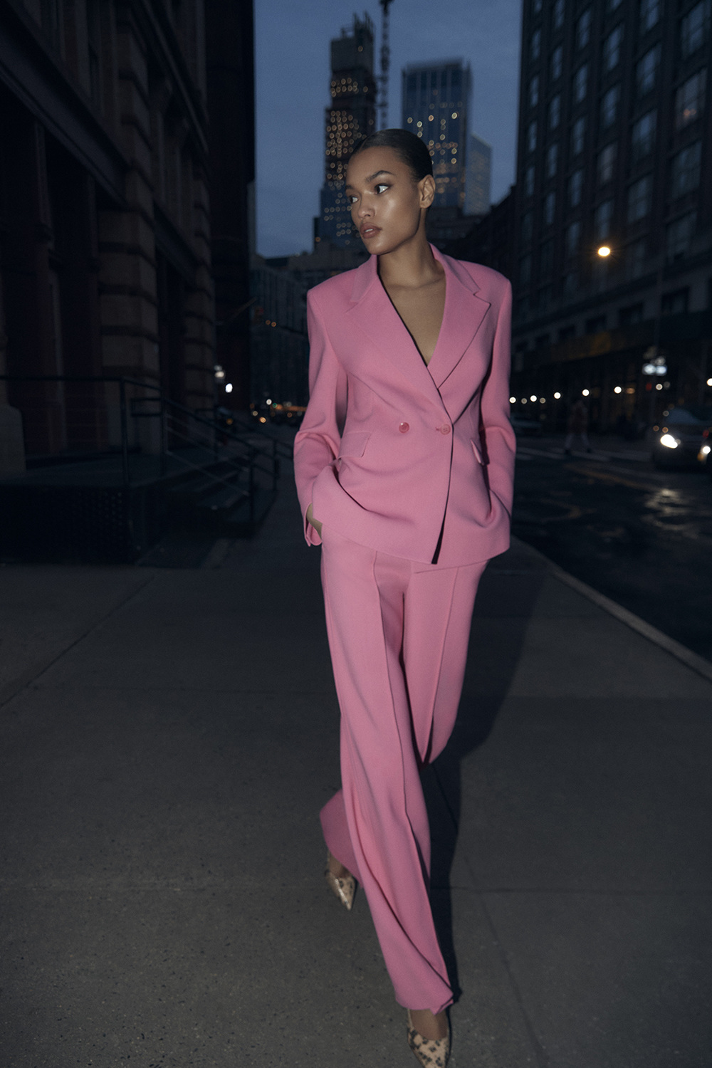 Elegant adult woman in a pink pantsuit confidently walks along a city street at dusk in front of tall buildings in an outdoor shot.