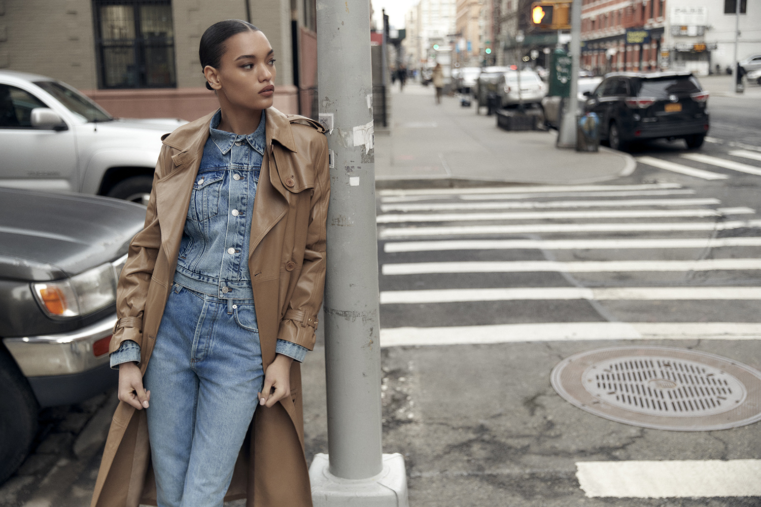 A woman in a trench coat stands on a city street next to a gray pole, wearing a denim jacket and jeans, with cars in the background.