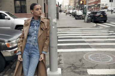 Model Lameka Fox leaning against a pole on a city street wearing a tan leather trench coat over a denim jacket and jeans.