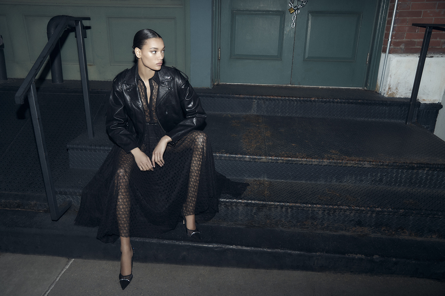 Model Lameka Fox seated on metal steps wearing a black leather jacket and sheer lace dress for Scanlan Theodore
