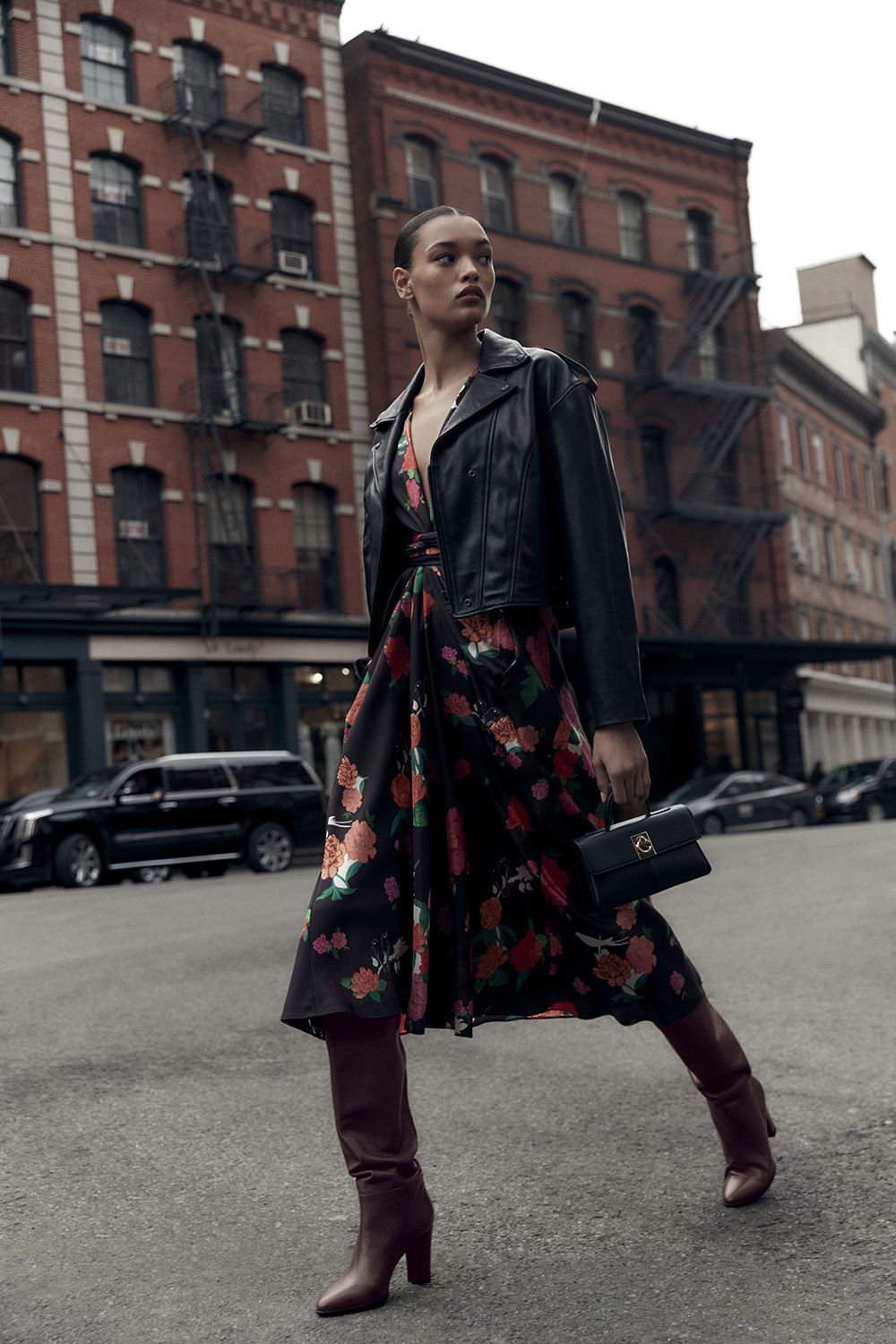 Fashionable woman in floral dress, leather jacket, and boots walking on urban street with brick buildings and cars in background.