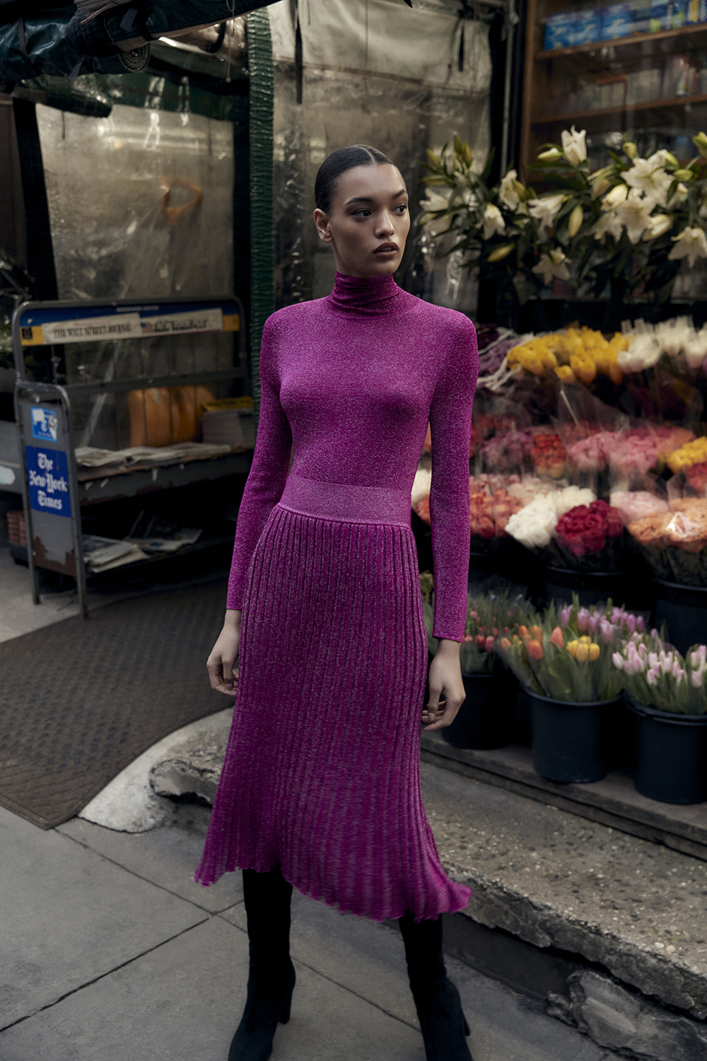 A woman stands outside a flower shop wearing a magenta turtleneck and pleated skirt with black boots on an overcast day in the city.