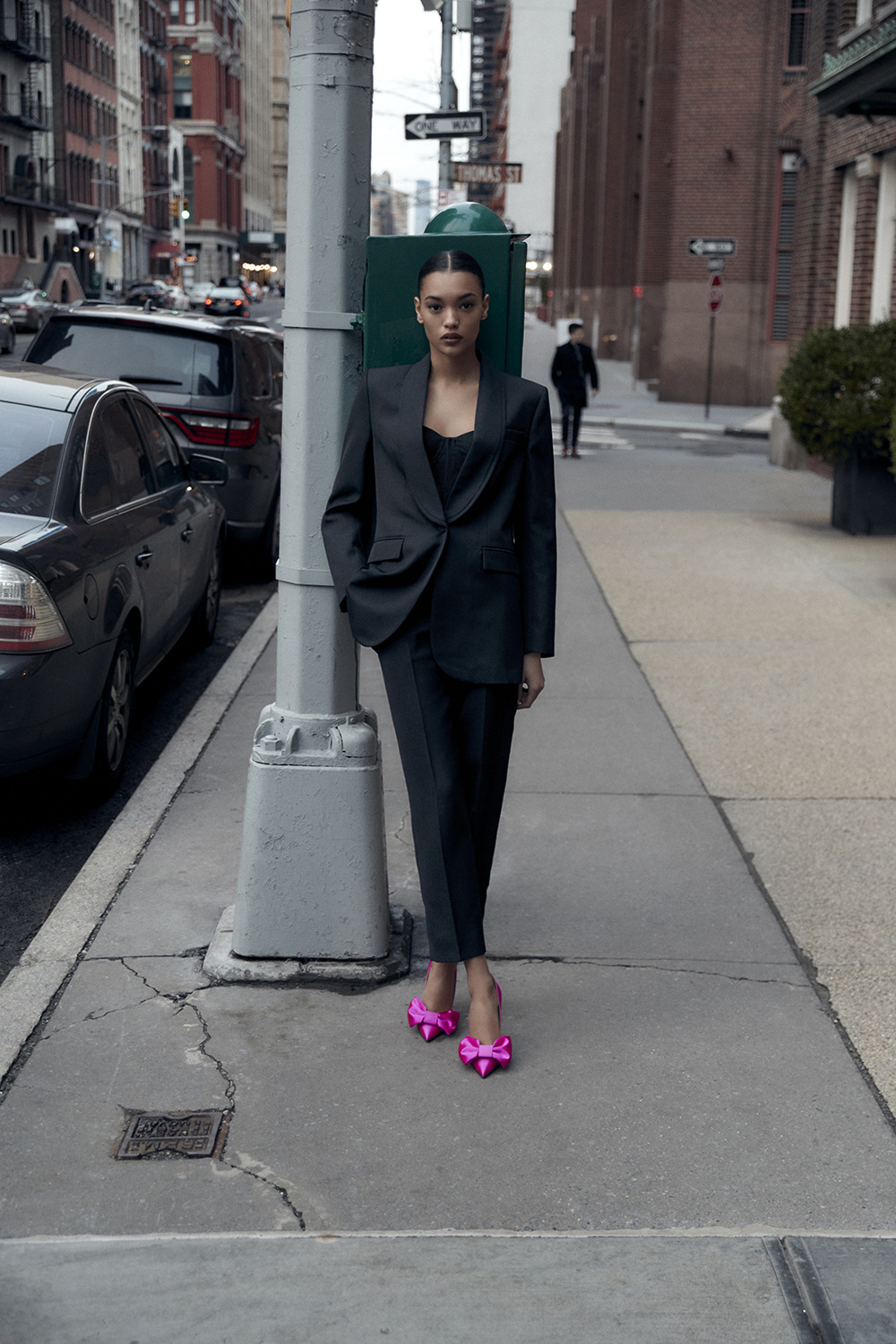 A woman in a dark suit stands on a sidewalk in an urban environment, posing with her hands in her pockets, with bright pink heels.
