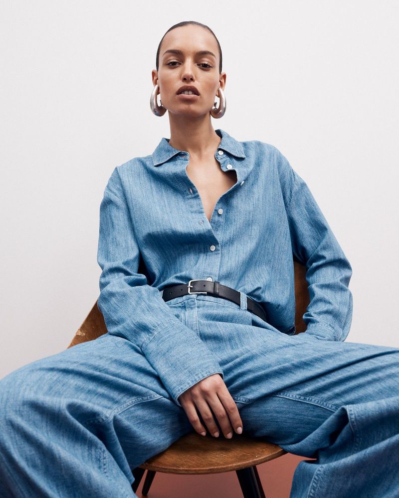 Fashion portrait of an adult woman in a denim shirt and pants, sitting in a chair against a white background, wearing silver hoop earrings.